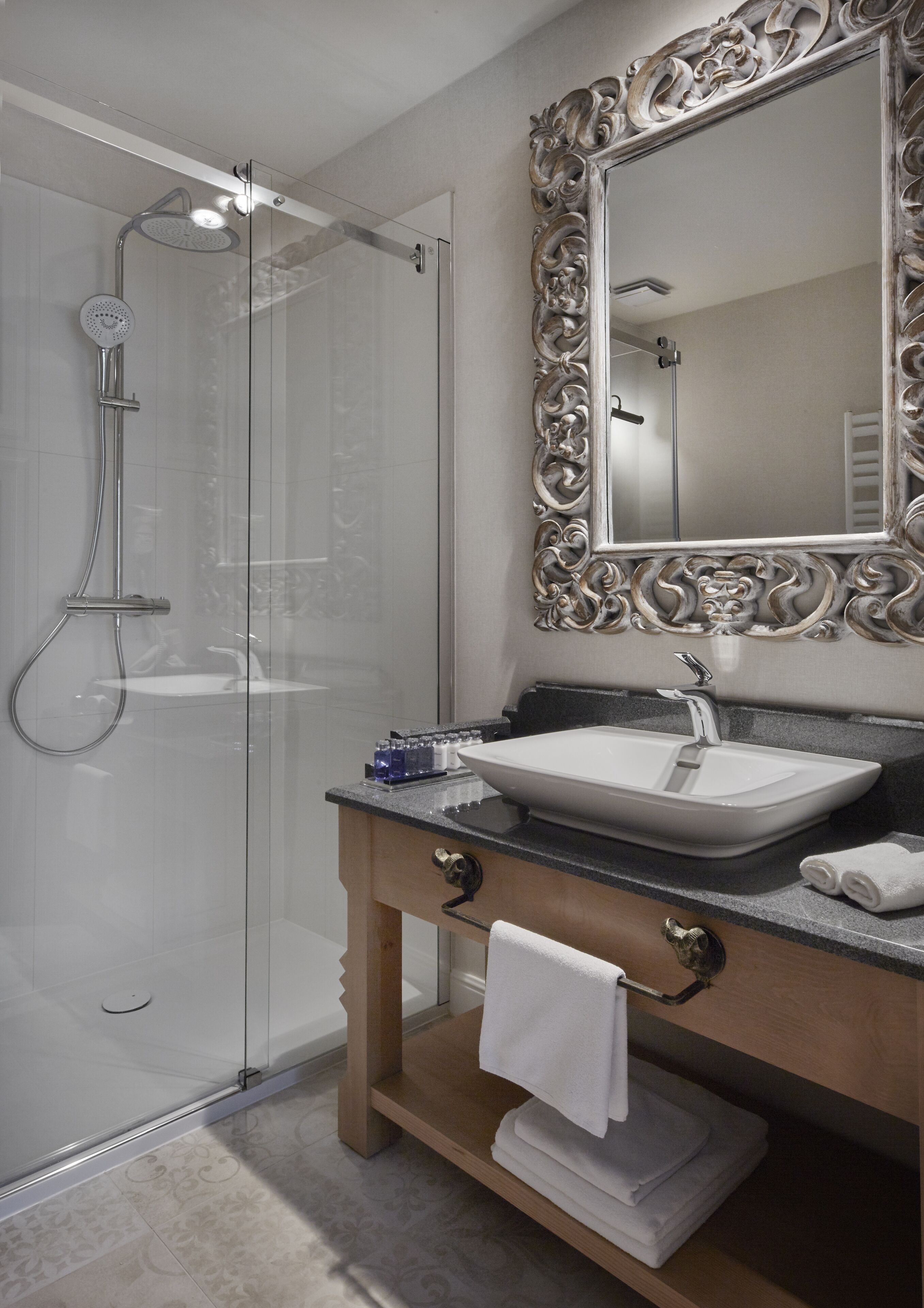 A modern bathroom with a glass-enclosed shower, a large ornate mirror, and a wooden vanity with a sink and towel rack.
