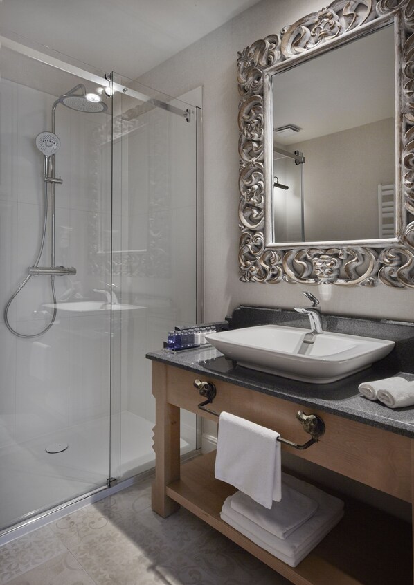Un baño moderno con ducha envuelta en vidrio, un espejo grande y ornamentado, y un tocador de madera con lavamanos y perchero para toallas.