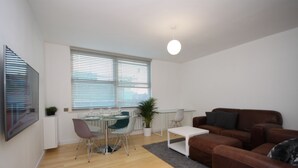 Ein modernes Wohnzimmer mit einer braunen Ledersofa, einem Glastisch mit Stühlen, einer Zimmerpflanze und einem großen Fenster mit Jalousien.