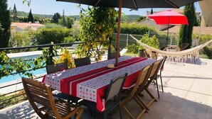 Um pátio externo com uma mesa posta para dois, rodeado por cadeiras, sob um grande guarda-sol.