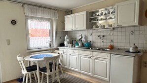 A compact kitchen with white cabinets, a small dining area with two chairs, and a window with lace curtains. - Vacation home 05 Juliane - W1 (Esens)