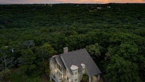 Una casa grande rodeada de un bosque frondoso, con un porche visible y un pequeño jardín.