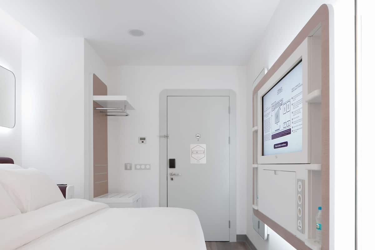 Une chambre d’hôtel avec un lit, une table de chevet, un téléviseur fixé au mur, et une porte dotée d’une serrure et d’un trou de serrure.