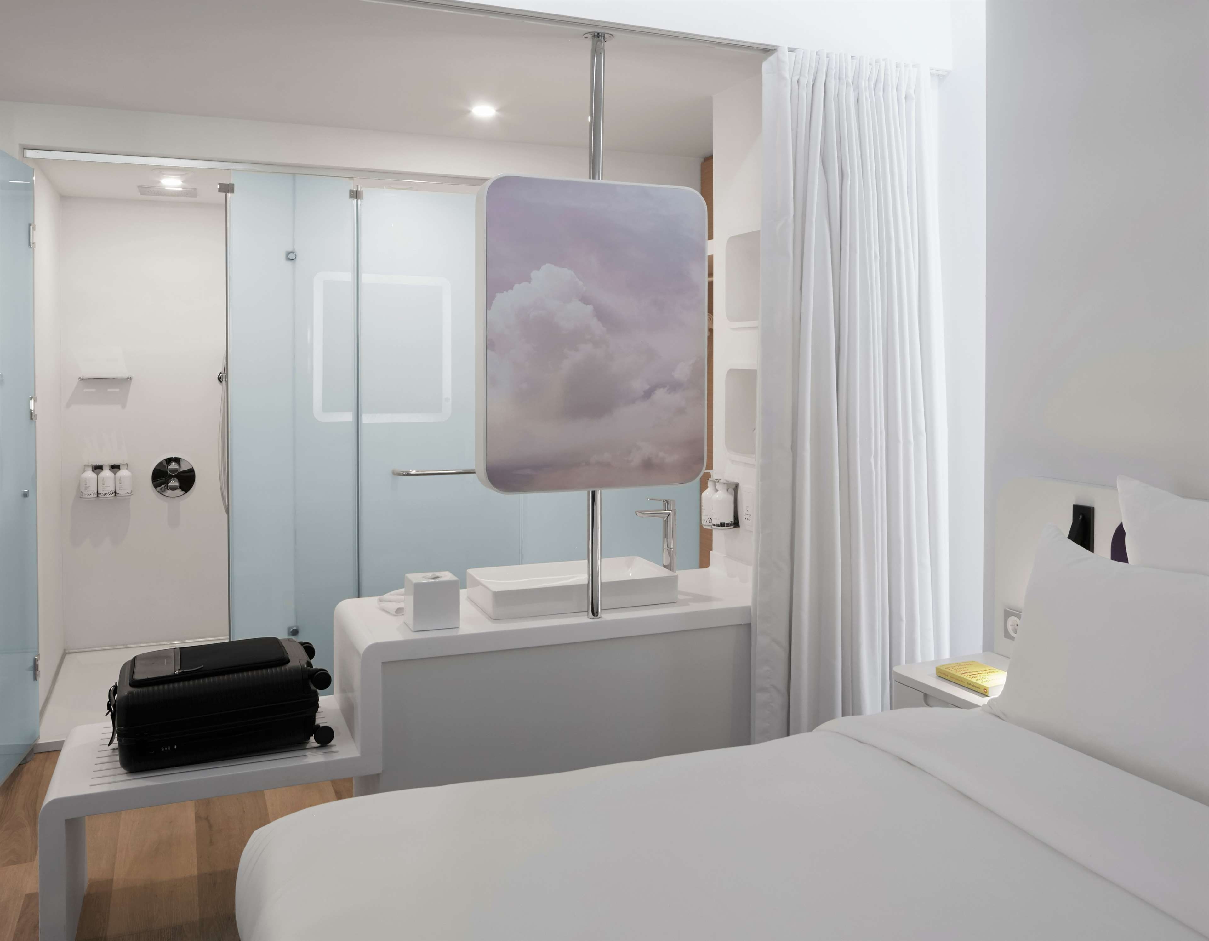 A hotel room with a bed, a bedside table, a desk with a computer, a window with curtains, and a bathroom with a shower and toilet.