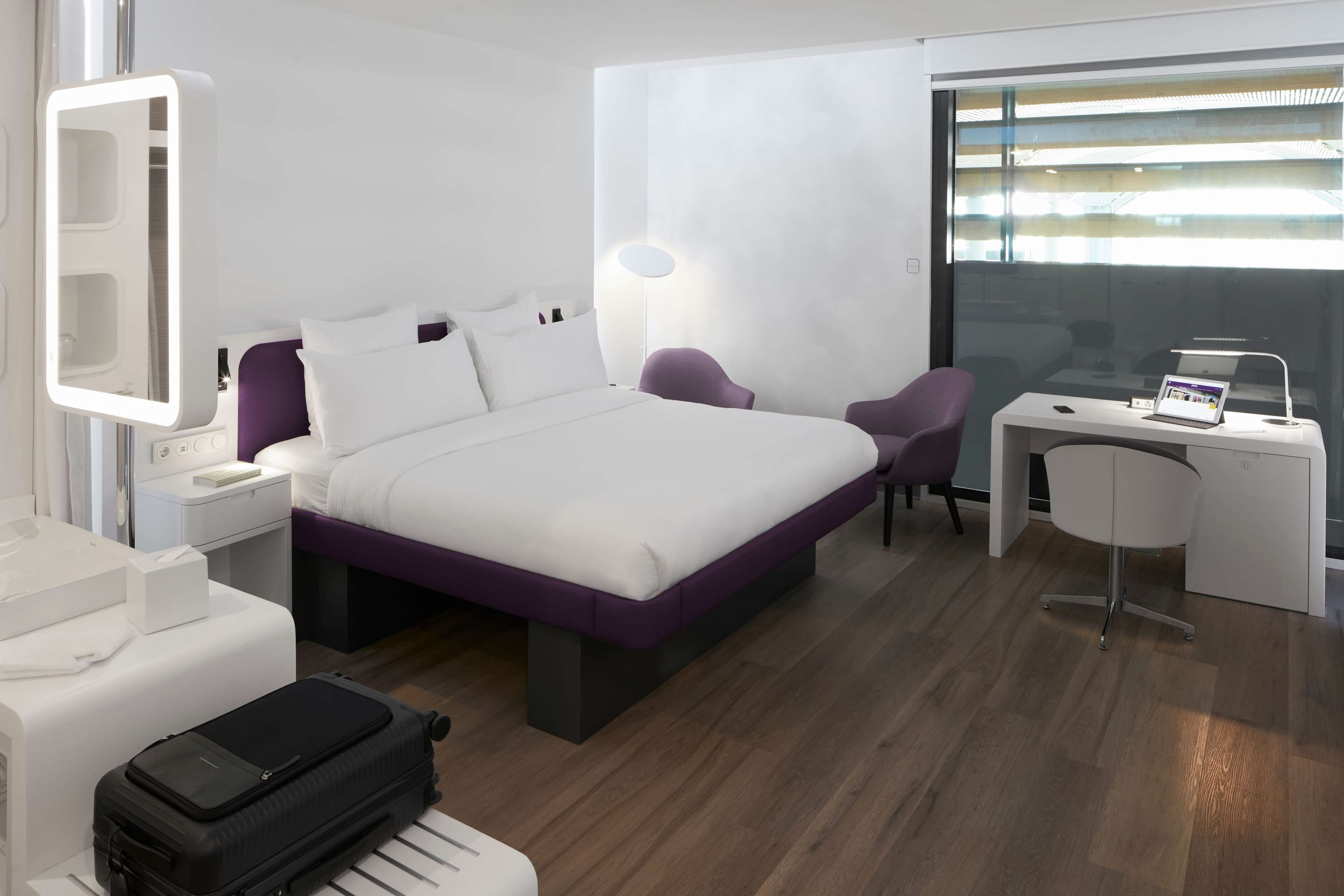 A modern hotel room with a large bed, a desk, a chair, a suitcase, and a mirror.