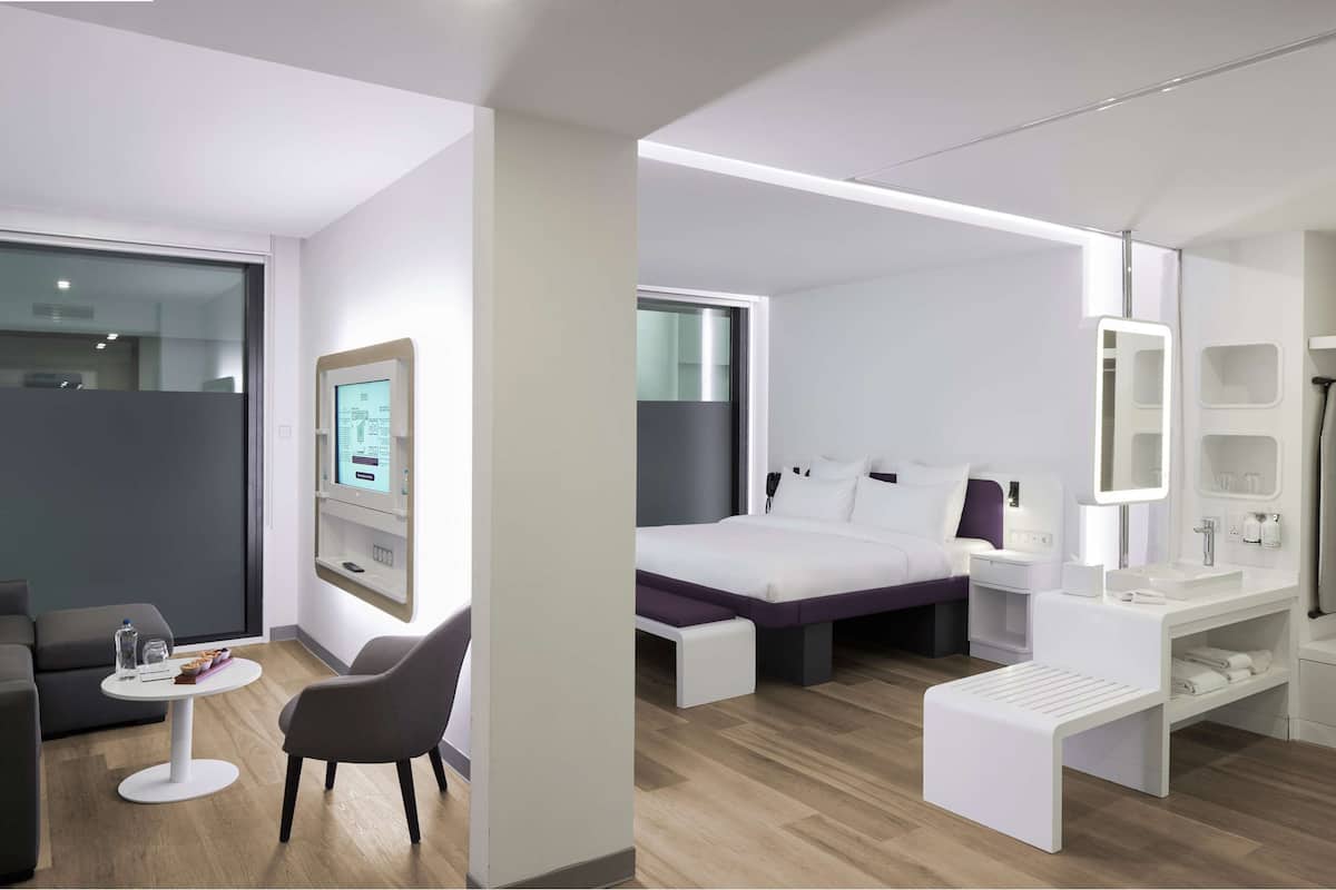 Une chambre d’hôtel moderne équipée d’un canapé, d’une petite table ronde, d’un lit avec une table de chevet, d’une coiffeuse avec un miroir et d’un écran numérique fixé au mur.