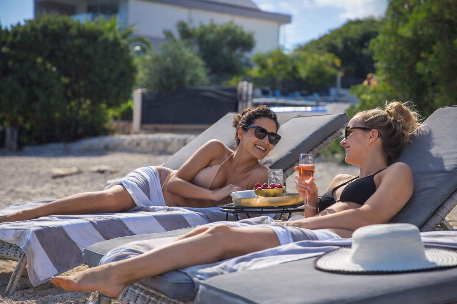 Two people relaxing on sun loungers with a table between them, a hat, and a drink.
