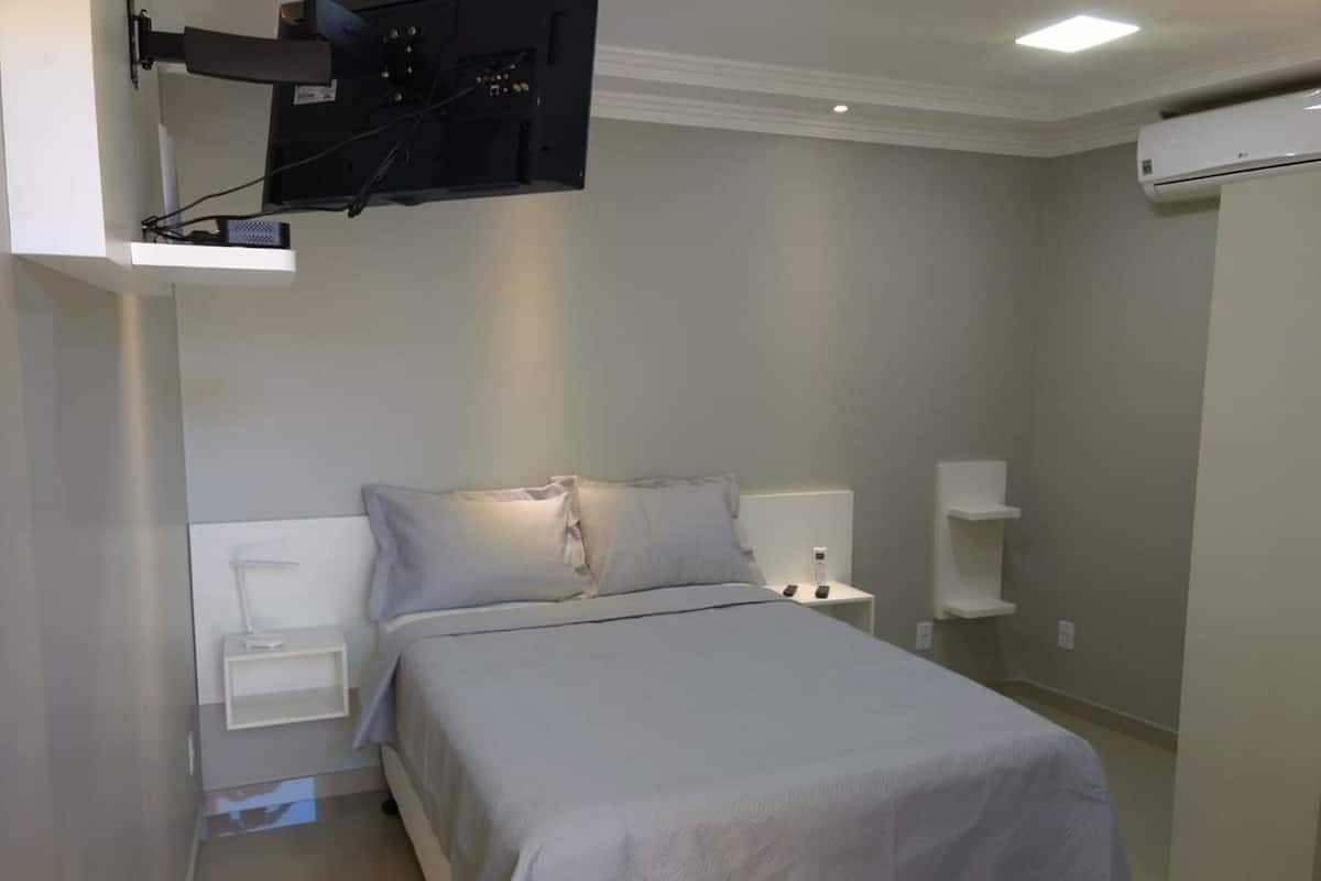 Quarto com cama, mesas de cabeceira, TV fixada na parede e ar condicionado.