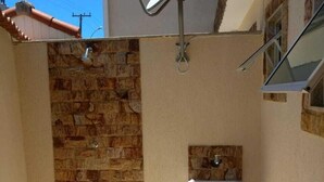 Un baño exterior pequeño y cerrado, con pared de piedra, un lavamanos blanco y zona de ducha.