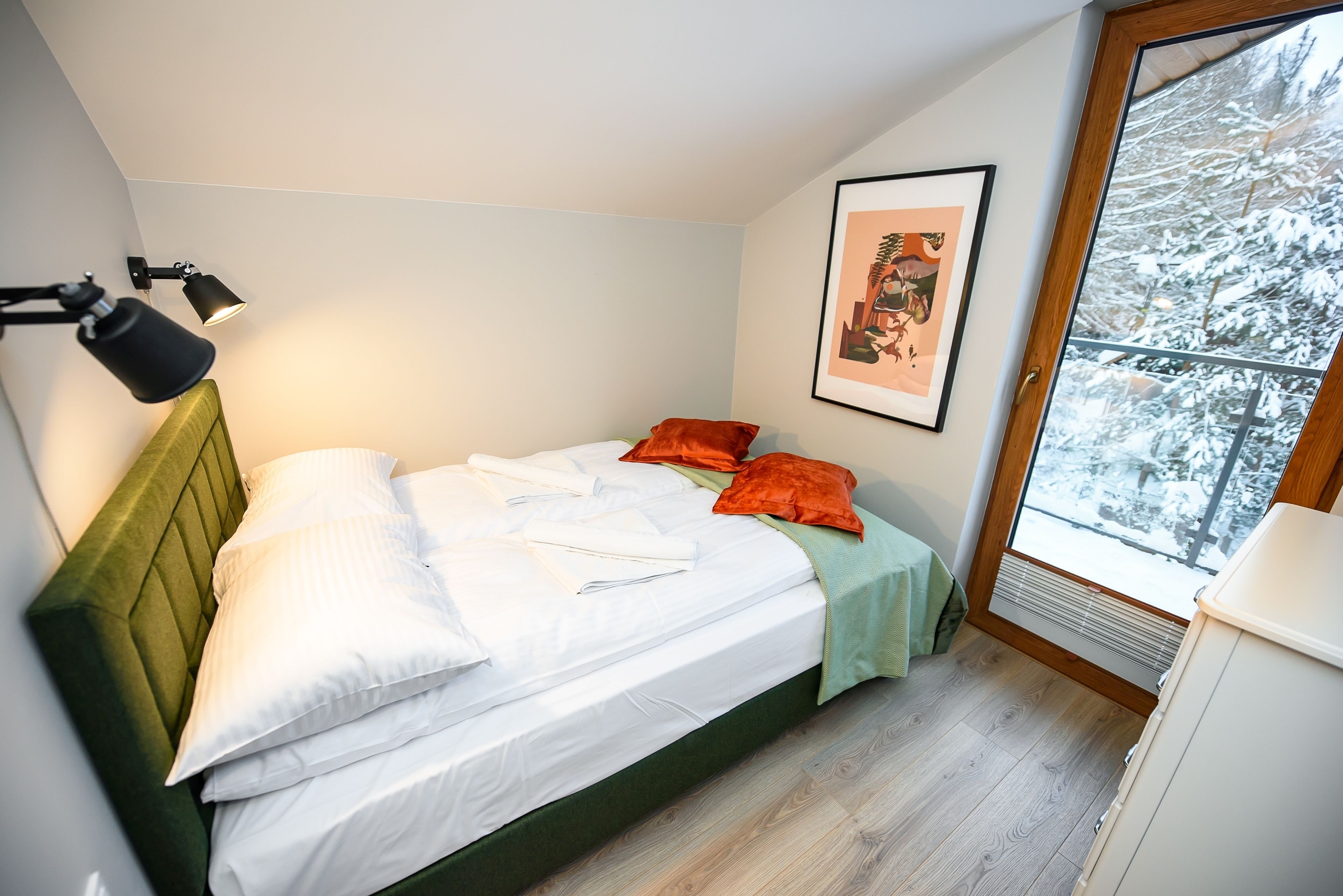 A neatly made bed with white linens and orange pillows, a green headboard, and a framed picture on the wall.