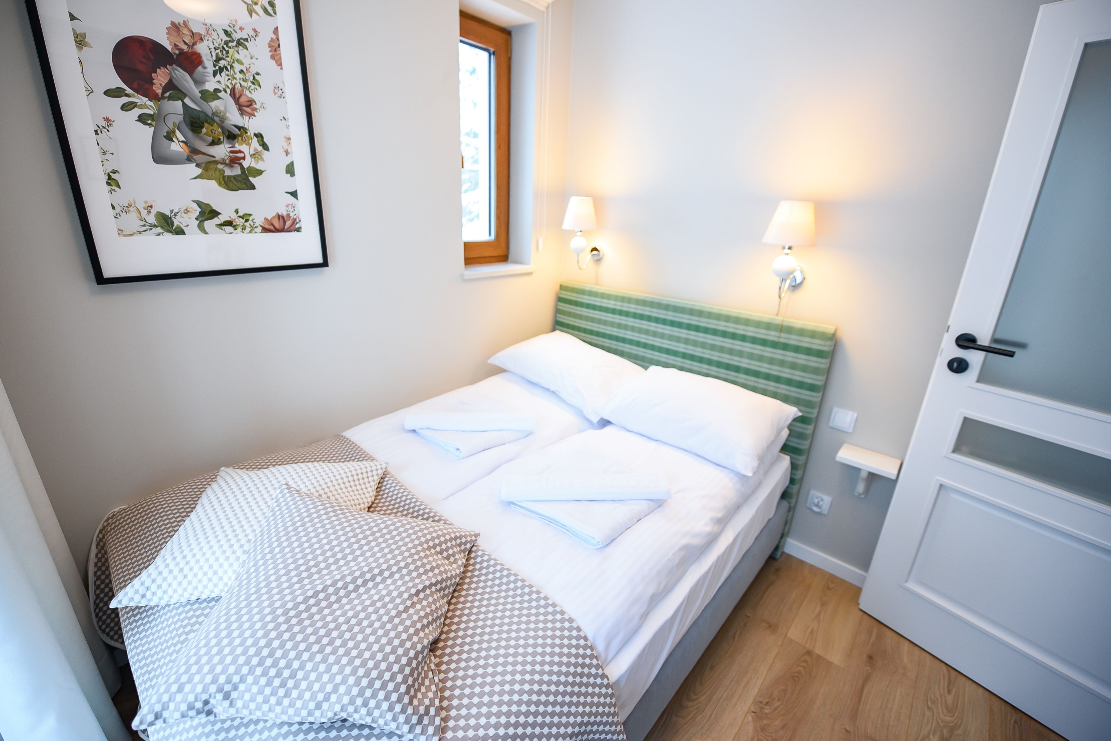 A neatly made bed with white linens and a green-striped headboard, two bedside lamps, and a framed floral artwork on the wall.