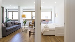 Una habitación de hotel moderna con un sofá gris, una pequeña mesa redonda, una silla, una cama con ropa blanca y ventanas grandes que dejan entrar luz natural.