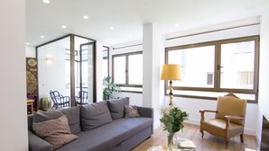 Una sala de estar moderna con un sofá gris, un sillón de madera y una pequeña mesa con una lámpara y libros.