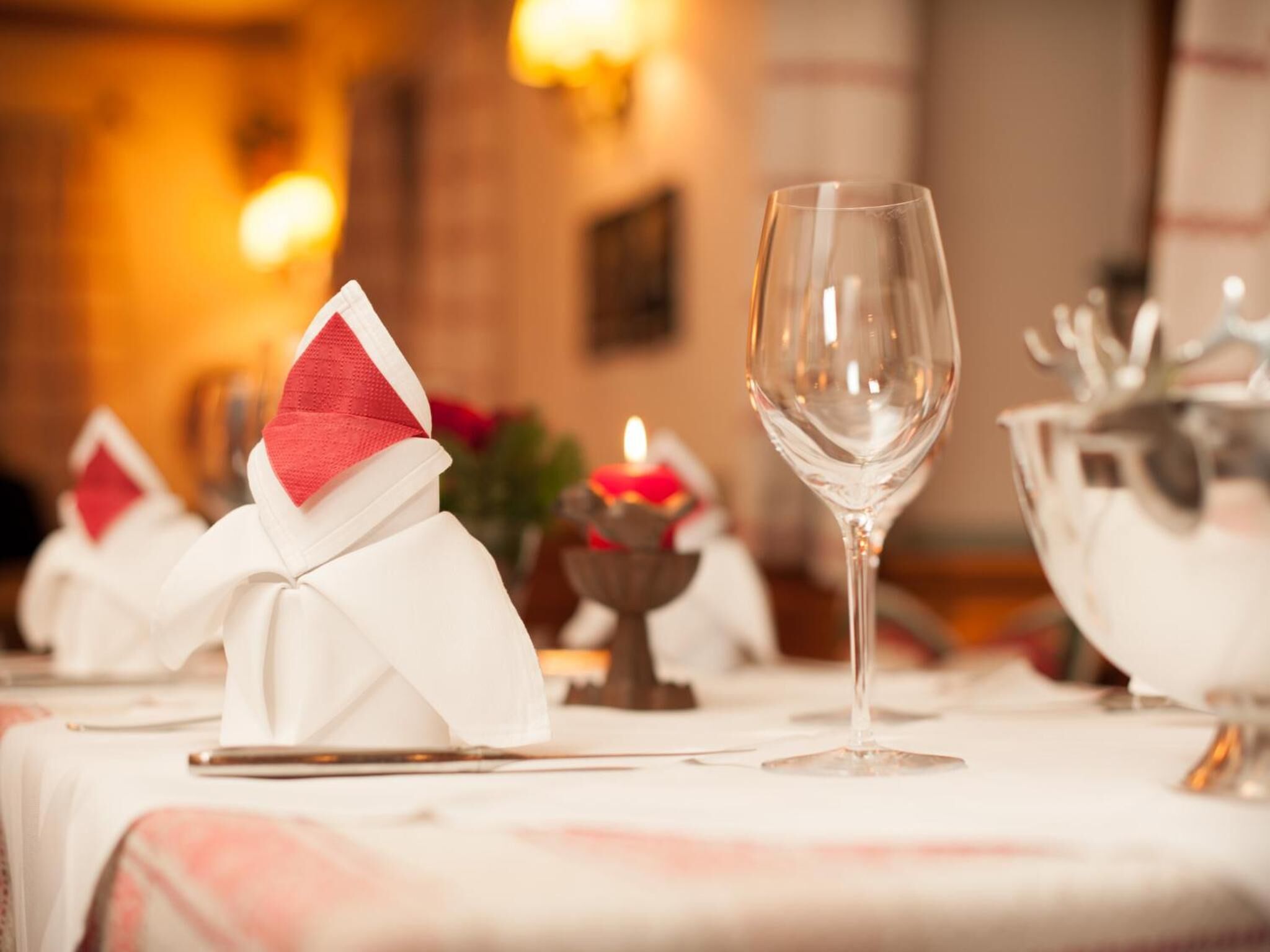 Una mesa de comedor dispuesta con servilletas blancas atadas con cintas rojas, una copa de vino transparente y una vela en un candelabro.