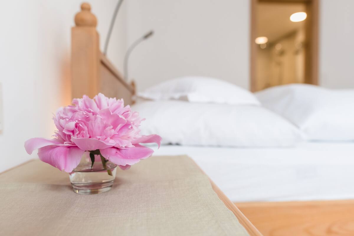 Ein Bett mit weißer Bettwäsche und ein kleiner Vasen mit rosa Blumen auf einem Nachttisch.