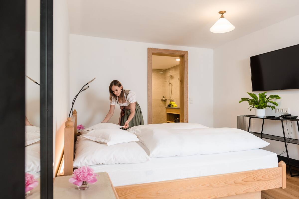 Ein ordentlich bezogenes Bett mit weißen Laken, ein Holzkopfteil und ein Nachttisch mit einer Vase voller rosa Blumen. Ein Fernseher ist an der Wand befestigt, und eine Topfpflanze steht auf einem kleinen Tisch neben dem Bett.