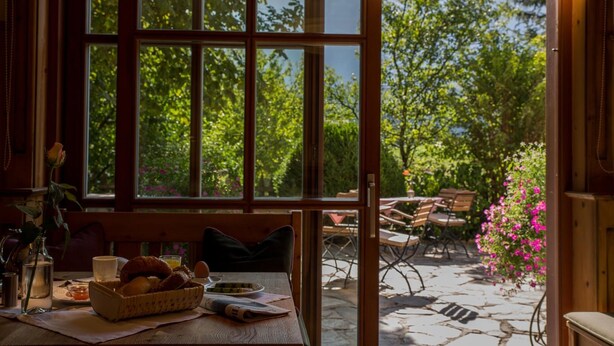 Ein gemütlicher Essbereich mit einem für das Frühstück gedecktem Tisch, einem Fenster mit Blick auf Grünflächen und einer Terrasse mit Sitzgelegenheiten.