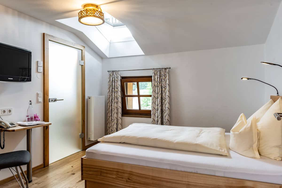 Ein Schlafzimmer mit Bett, Schreibtisch mit Stuhl, Wand-Fernseher, Fenster mit Vorhängen und einem Oberlicht.