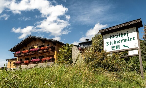 Ein traditionelles Holzhaus mit Balkonen, die mit Blumen geschmückt sind, und einem Schild mit der Aufschrift 'Gasthof Steinert', eingebettet in eine natürliche, verwilderte Umgebung.