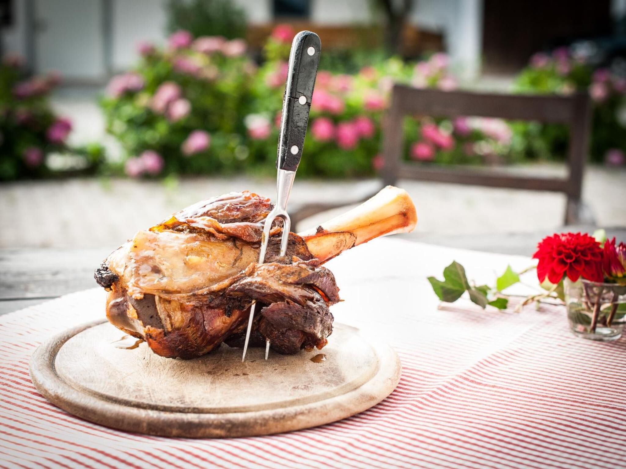 A roasted lamb leg on a wooden board with a knife inserted, placed on a table with a red and white striped tablecloth.