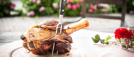 A roasted lamb leg on a wooden board with a knife inserted, placed on a table with a red and white striped tablecloth.