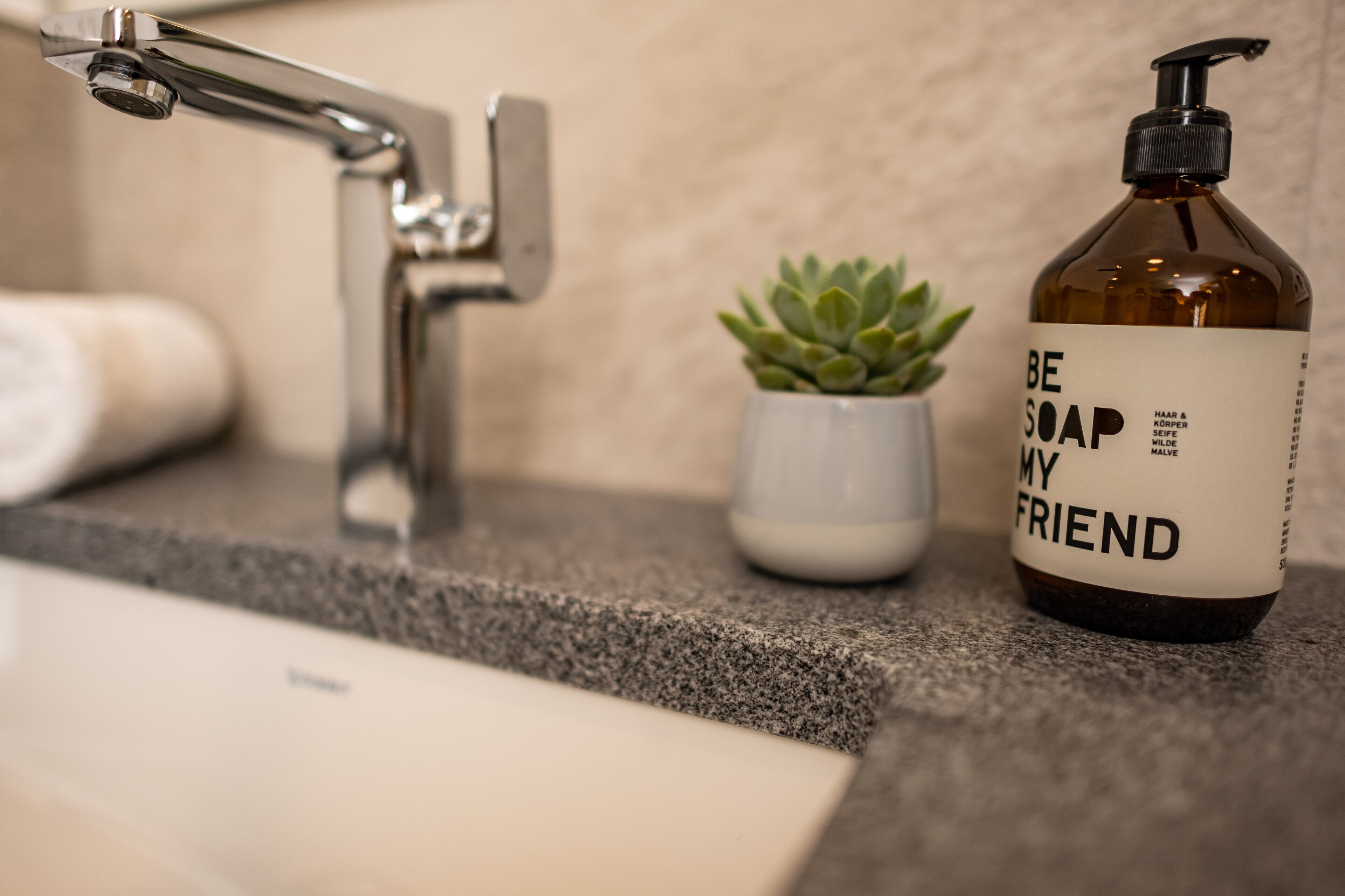 A bathroom with a granite countertop, a faucet, a small potted plant, and a bottle of soap.