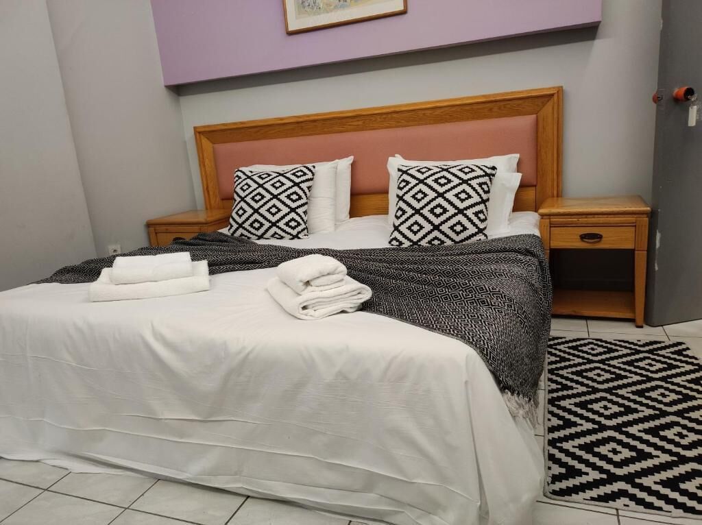 A neatly made bed with a white bedspread and patterned pillows, a wooden headboard, a bedside table with a lamp, and a patterned rug on the floor.