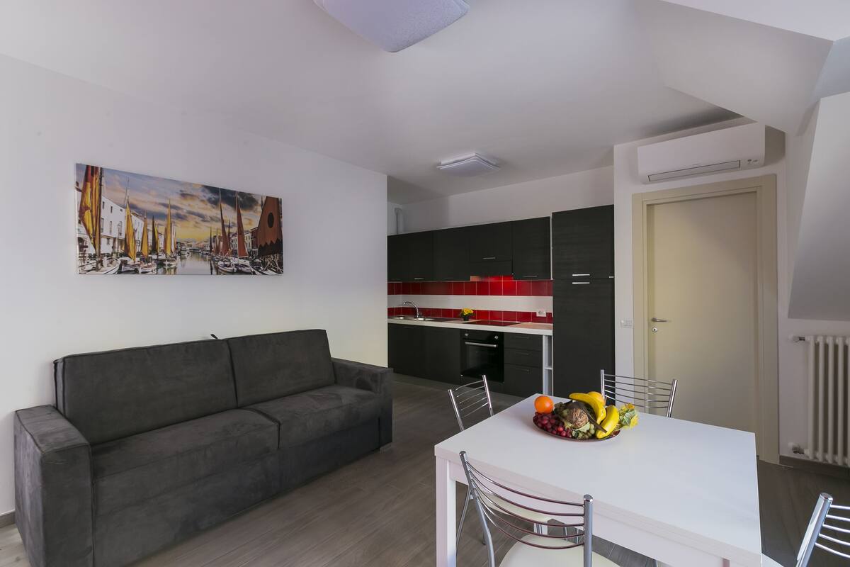A modern living space with a grey sofa, a dining table with chairs, and a kitchen area with black cabinets and a red backsplash.