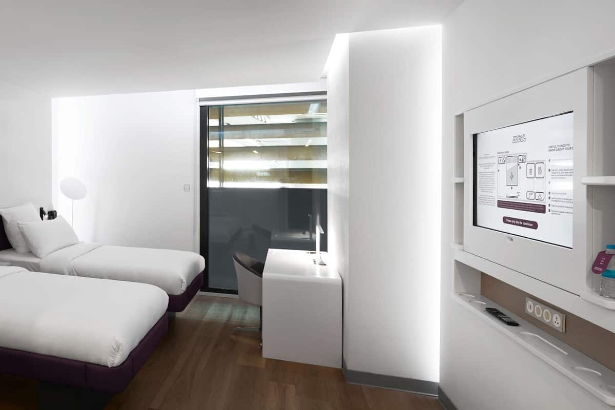 Une chambre d’hôtel moderne, dotée d’un grand miroir, d’une table de chevet avec une lampe, d’un lit avec une literie blanche et d’un petit bureau avec un ordinateur.