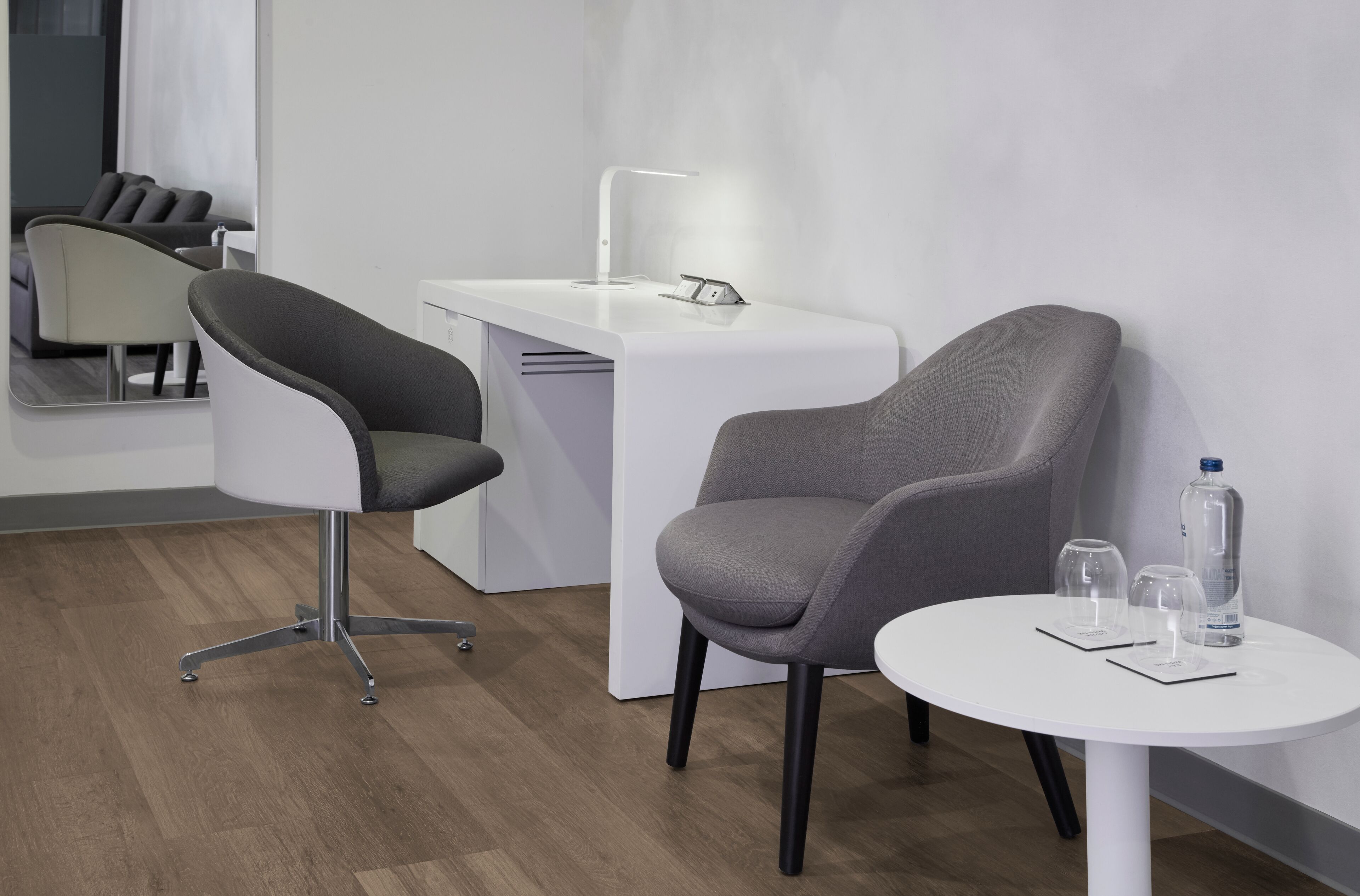 A modern hotel room with a white desk, a grey armchair, a round white table with a bottle and glasses, and a mirror reflecting the room's interior.