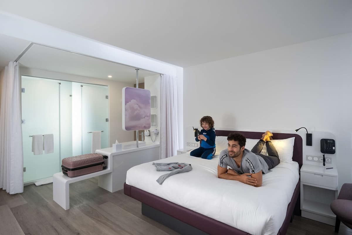 Une chambre d’hôtel moderne dotée d’un grand lit, d’une table de chevet, d’une petite table et d’une salle de bain visible à travers une paroi en verre.