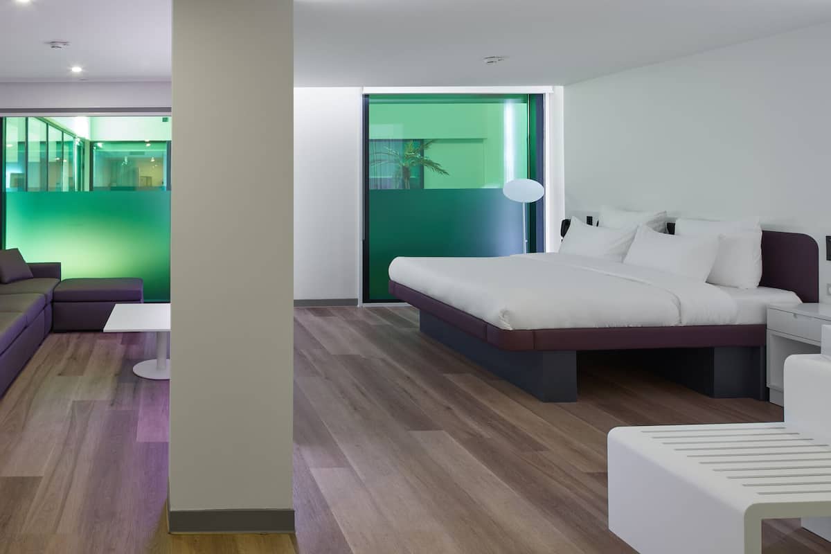 Une chambre d’hôtel moderne, équipée d’un canapé, d’une petite table ronde, d’un lit avec une tête de lit, d’une table de chevet et d’un miroir.