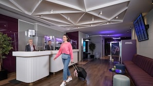 Een moderne hotellobby met een receptiebalie, een digitaal informatiebord, een zithoek en een houten vloer.