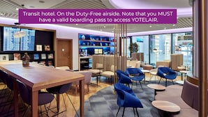 Een moderne hotellobby met een houten receptiebalie, een zithoek met blauwe stoelen en een digitaal displaybord.