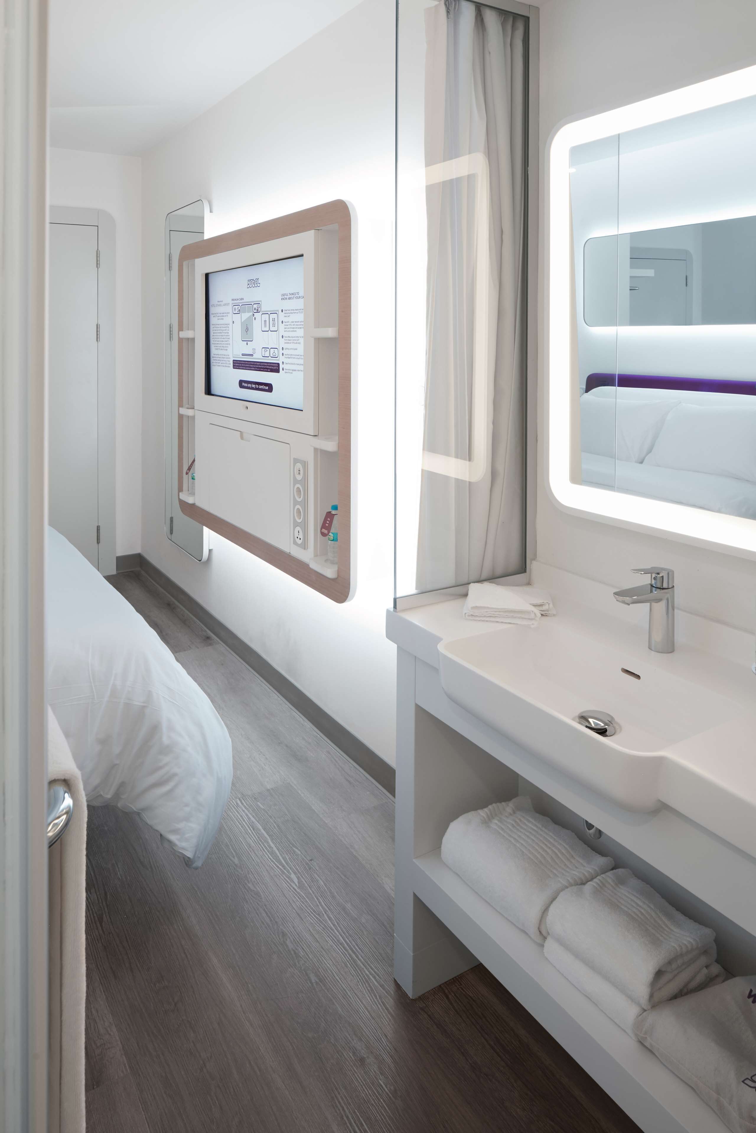 A hotel room with a bed, a bedside table with a mirror, a wall-mounted control panel, and a window with curtains.