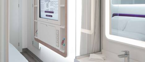 A hotel room with a bed, a bedside table with a mirror, a wall-mounted control panel, and a window with curtains.