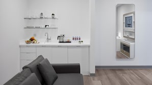 A modern kitchen with white cabinetry, a built-in oven, and a coffee maker. There is a small seating area with a grey sofa and a coffee table.