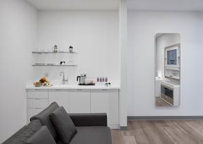 A modern kitchen with white cabinetry, a built-in oven, and a coffee maker. There is a small seating area with a grey sofa and a coffee table.