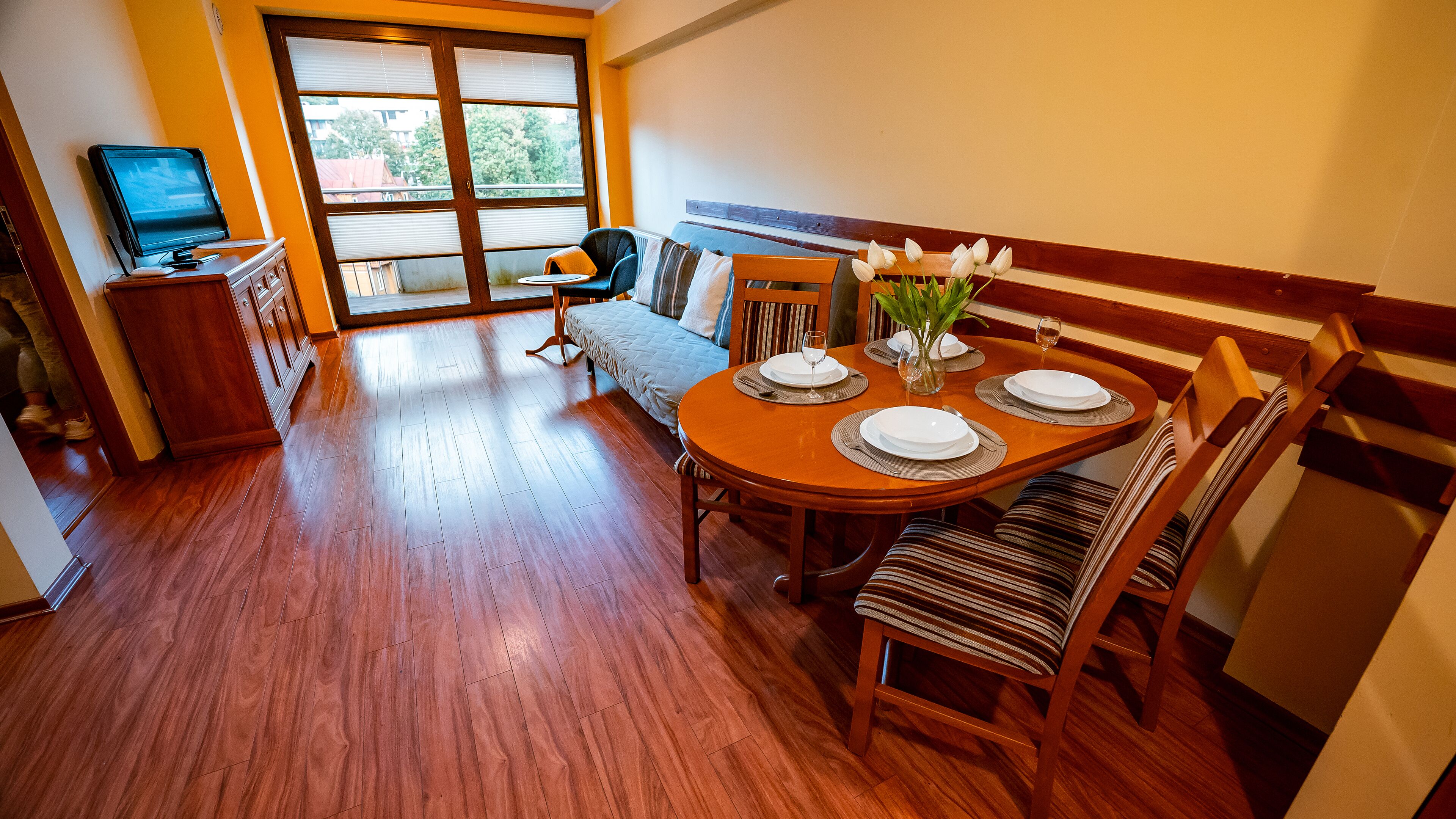 A dining area with a wooden table set for four, a bench, and a television in the corner.