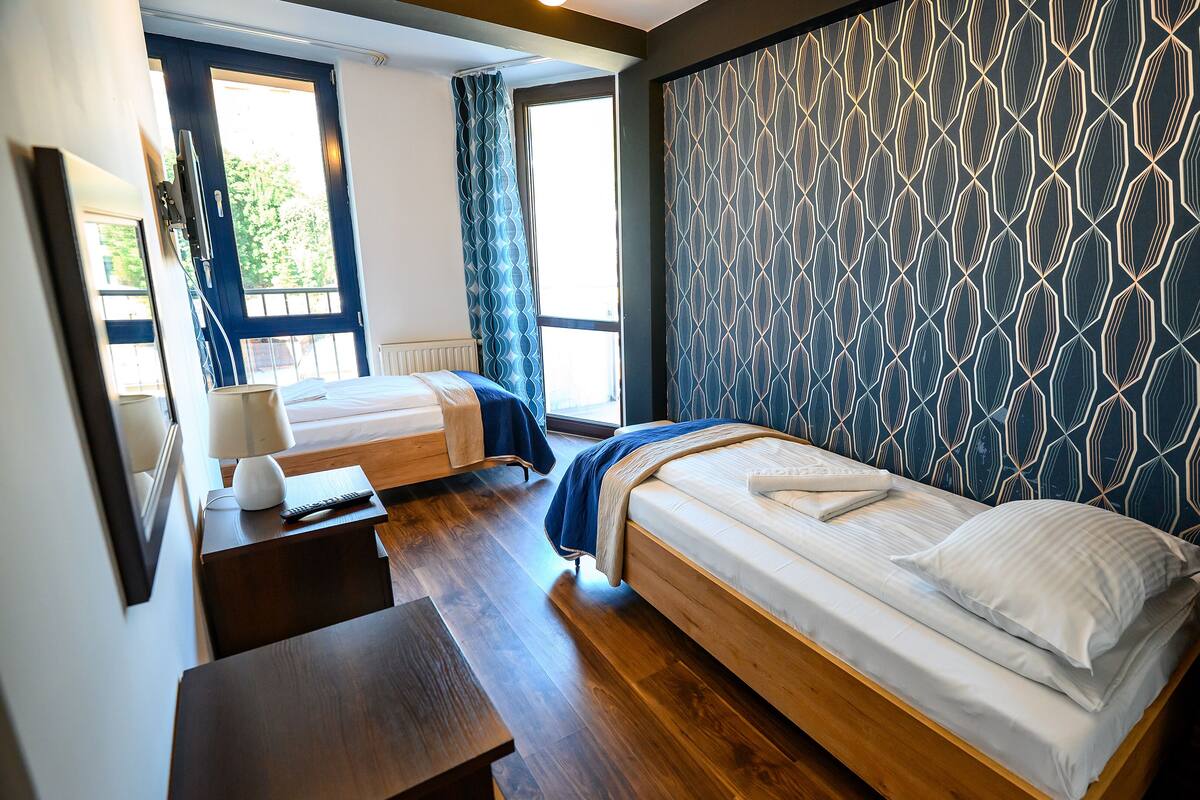 A hotel room with two single beds, a wooden desk, a chair, and a patterned wall.