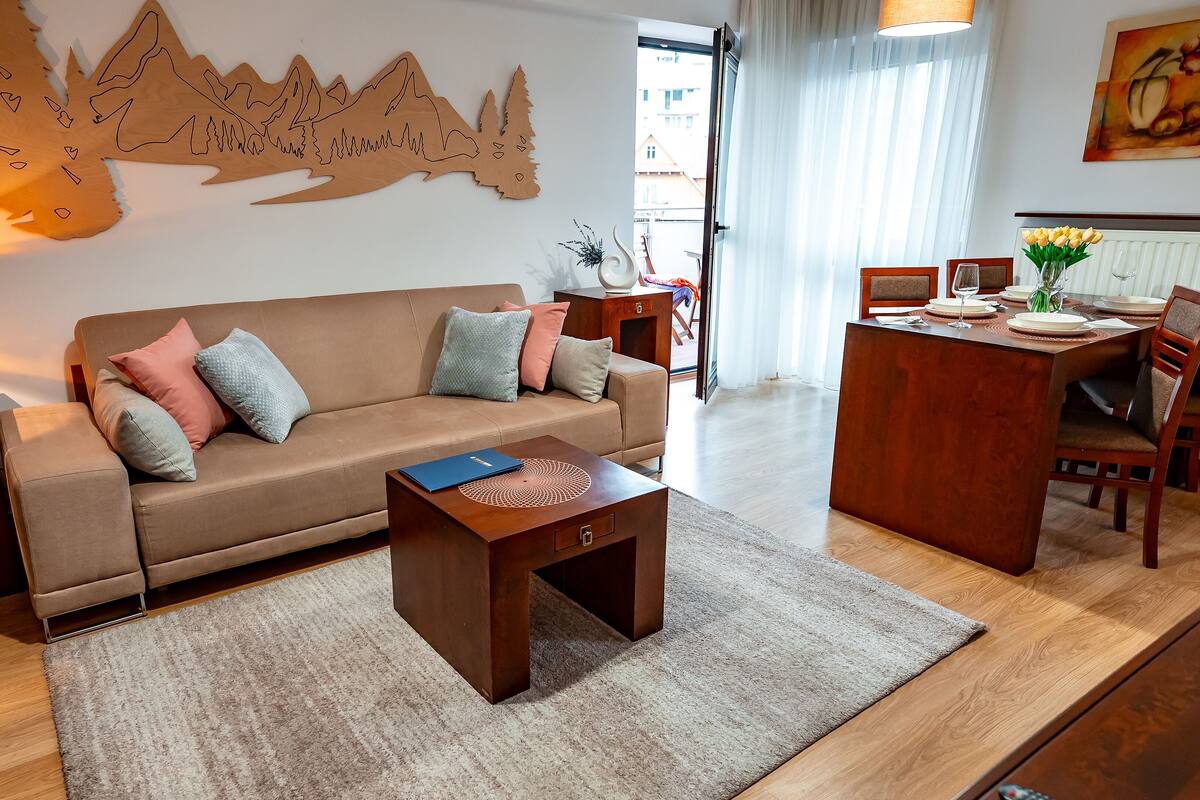 A living room with a sofa, coffee table, dining table with chairs, and a wall with a mountain design.