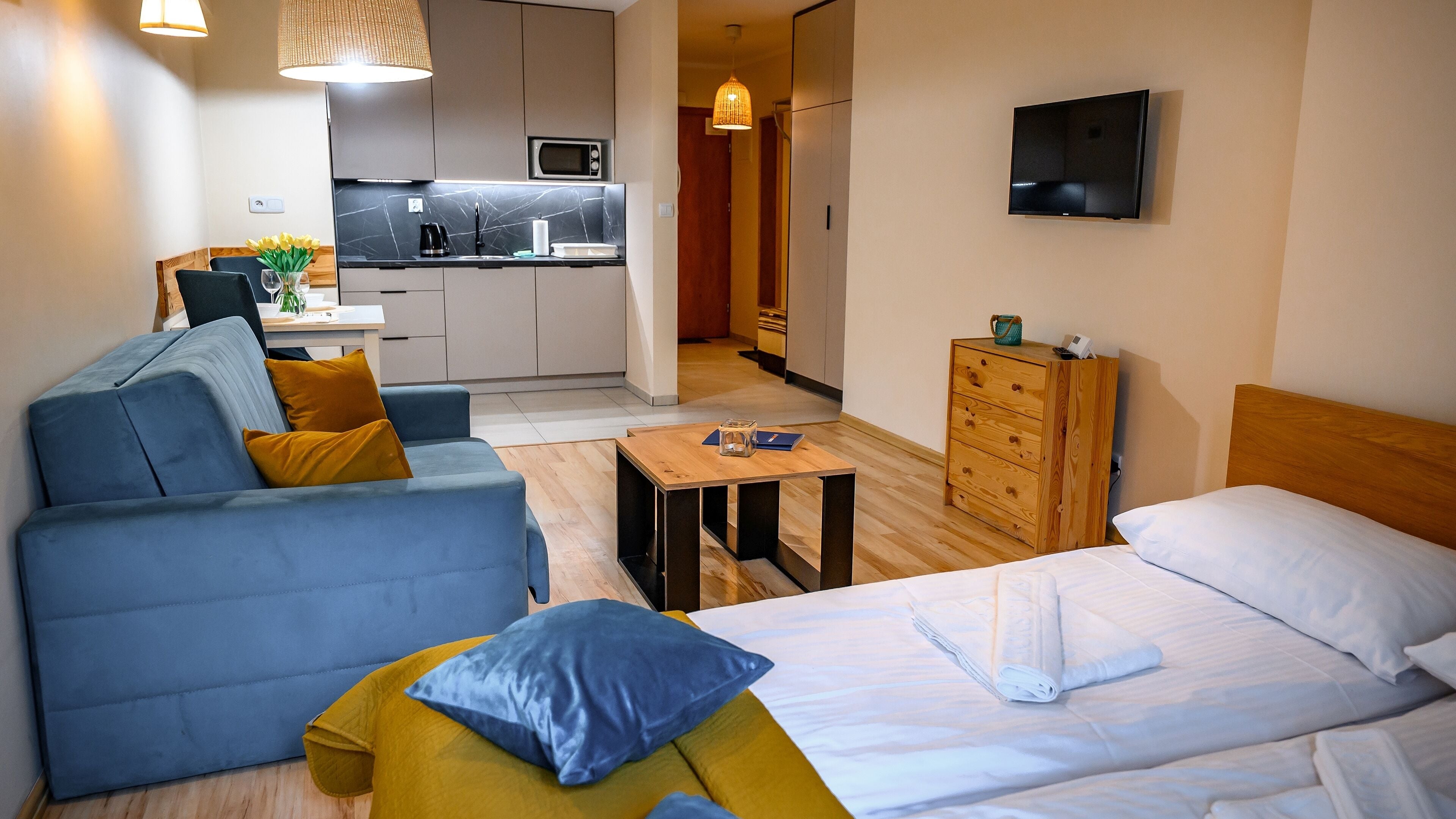 Ein modernes Hotelzimmer mit einer Couch, einem Bett, einem Holzkleiderschrank, einem kleinen Tisch und einer Küchenzeile im Hintergrund.