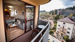 A balcony with a wooden floor, a grey sofa, a small table, and a chair. A large window offers a view of a building labeled "HOTEL KRYNICA" and surrounding landscape. - Sun & Snow Centrum Piłsudskiego (Krynica-Zdroj)