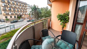 Un balcón con sillas de mimbre y una mesa de vidrio, con vista a una calle con coches aparcados y un edificio de varios pisos.