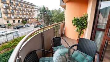 Un balcón con sillas de mimbre y una mesa de vidrio, con vista a una calle con coches aparcados y un edificio de varios pisos.