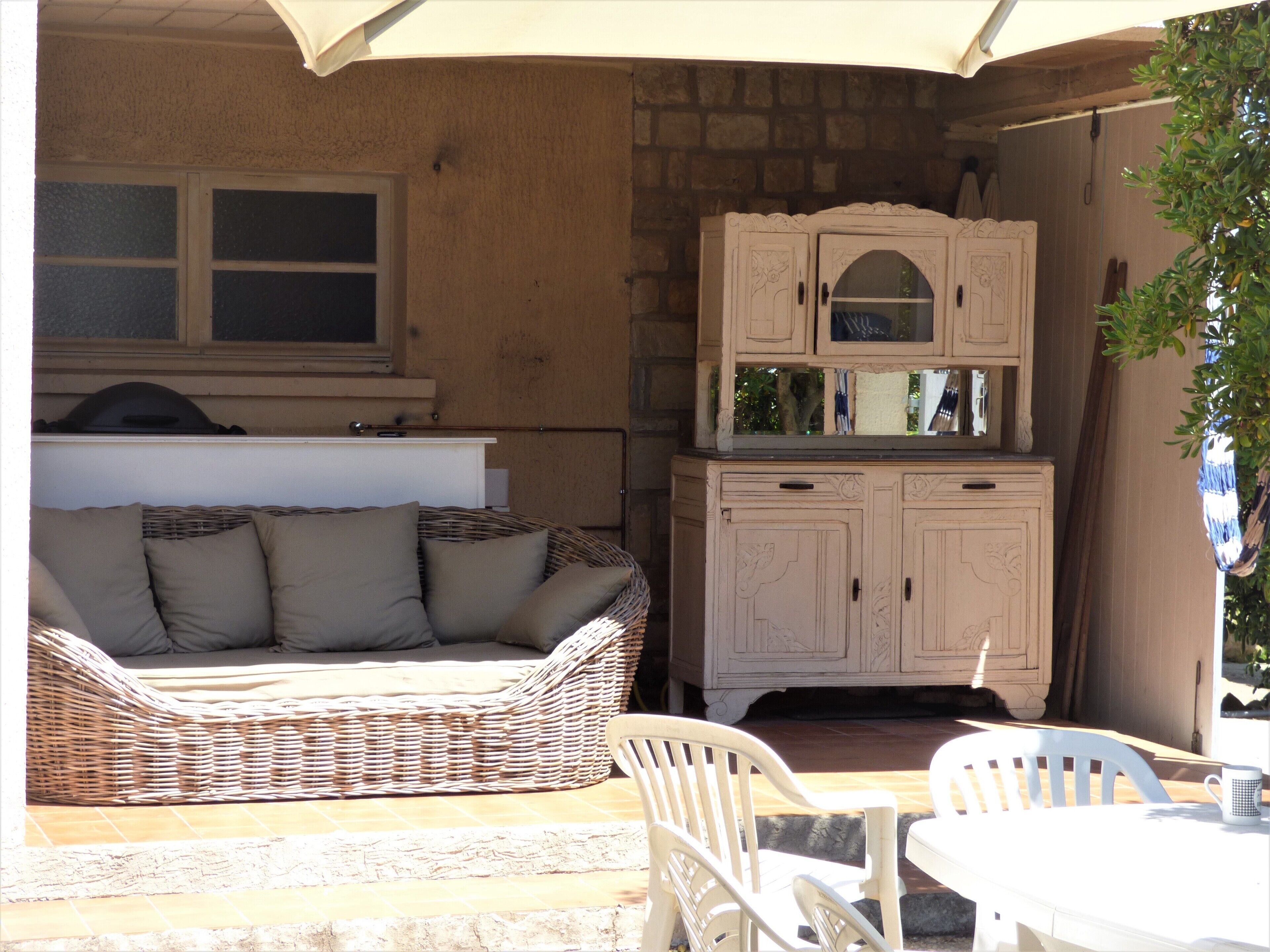 A wicker sofa with cushions, a white cabinet with decorative carvings, and a white table with chairs in a sunlit outdoor area.