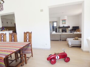 Un coin repas avec une table et des chaises en bois, une petite voiture rouge, une cuisine en arrière-plan et un salon avec un canapé et un téléviseur.