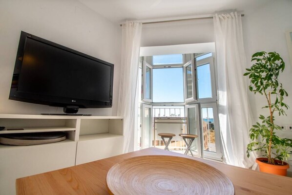 A modern room with a flat-screen TV on a white shelf, a wooden table in the foreground, a potted plant, and a large window with a view of the outside.