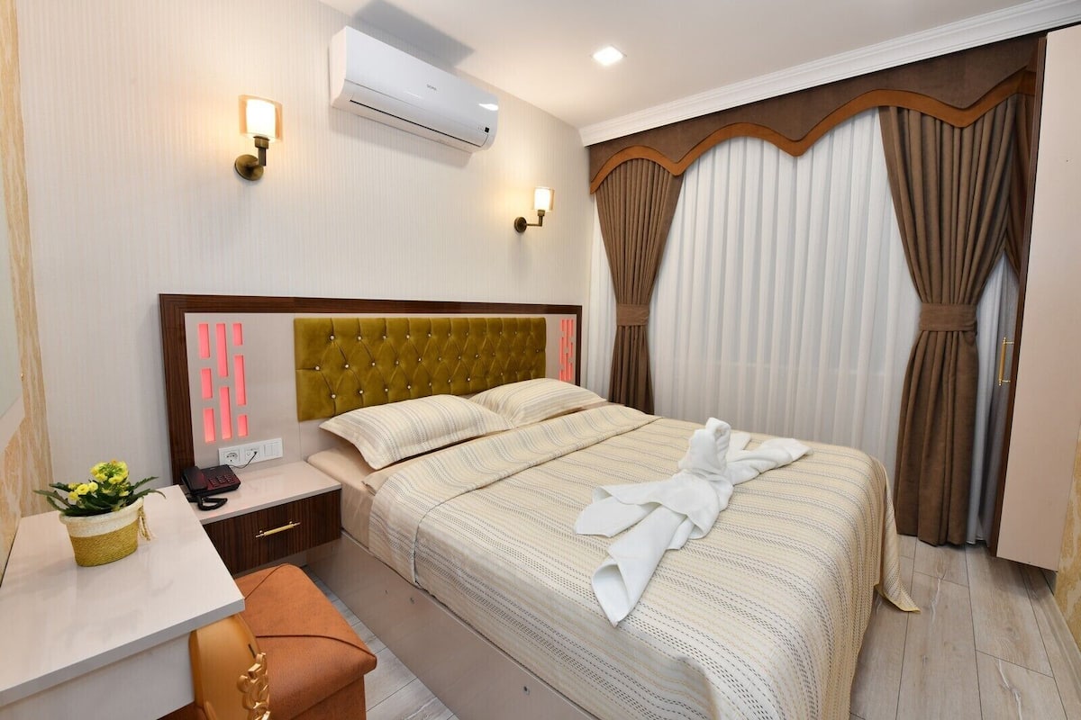 Habitación de hotel con cama, mesita de noche, lámpara, aire acondicionado, cortinas y una planta pequeña.