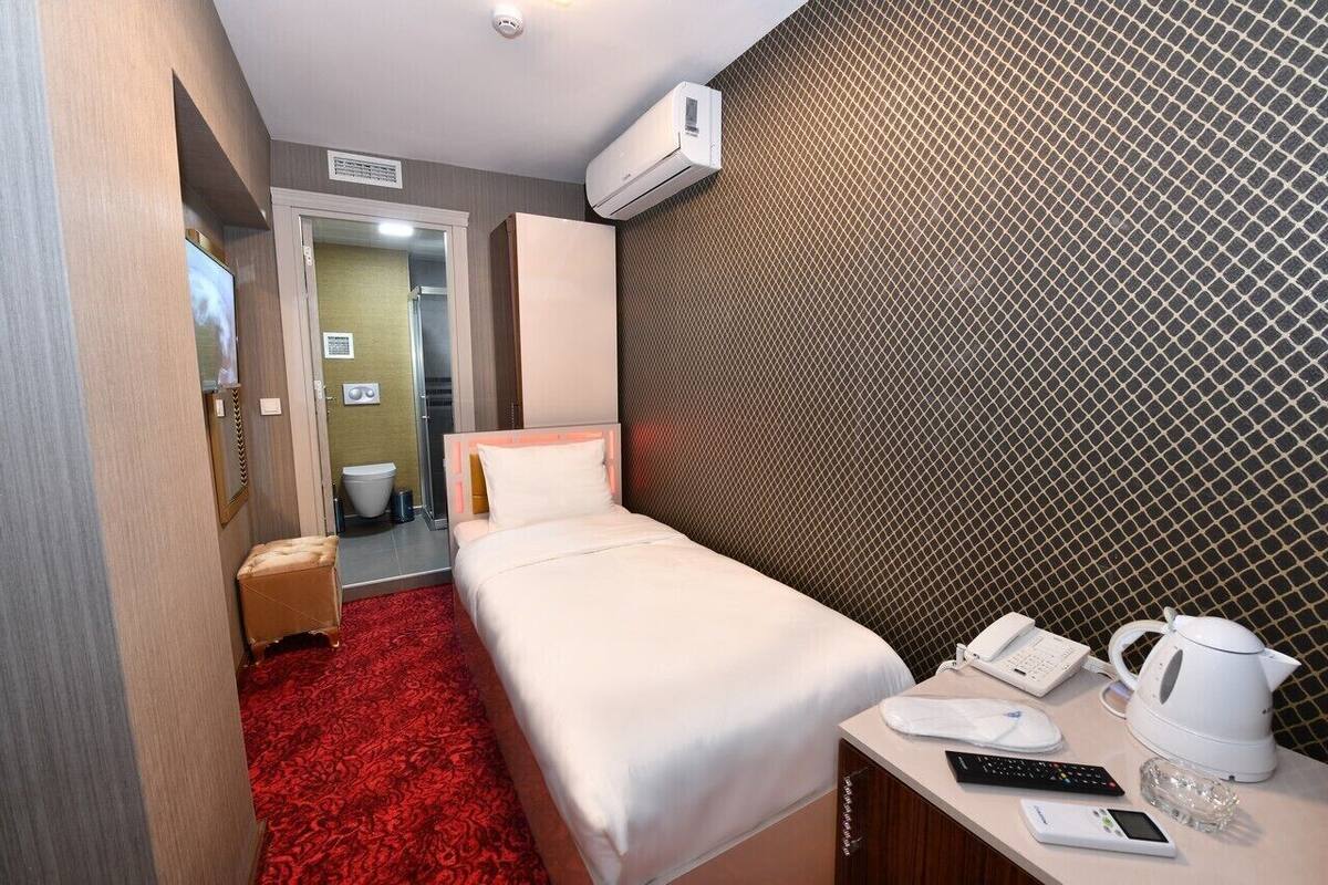 Habitación de hotel con una cama, alfombra roja, mesita de noche blanca con un teléfono, tetera y una pequeña mesa con un vaso.
